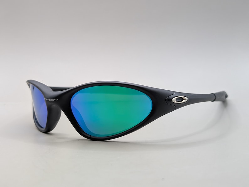 May include: Black sunglasses with a sleek, curved design. The lenses have a vibrant green and blue gradient. The frame features the Oakley logo on the temple. These sunglasses are designed for sports and outdoor activities.