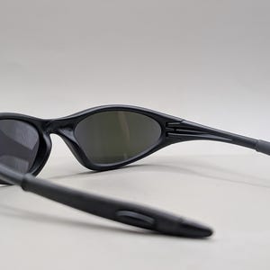 May include: Black sunglasses with dark lenses. The sunglasses have a sleek, curved design with a matte finish. The arms of the sunglasses are extended, showing the hinge and the ear pieces. The lenses are oval-shaped.