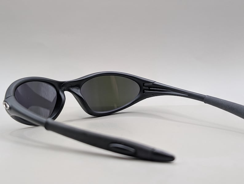 May include: Black sunglasses with dark lenses. The sunglasses have a sleek, curved design with a matte finish. The arms of the sunglasses are extended, showing the hinge and the ear pieces. The lenses are oval-shaped.