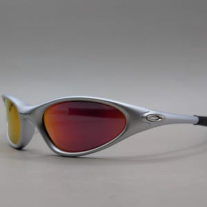 May include: Silver-framed sunglasses with red and yellow lenses. The sporty eyewear features a curved design and black temple tips. The Oakley logo is visible on the frame.