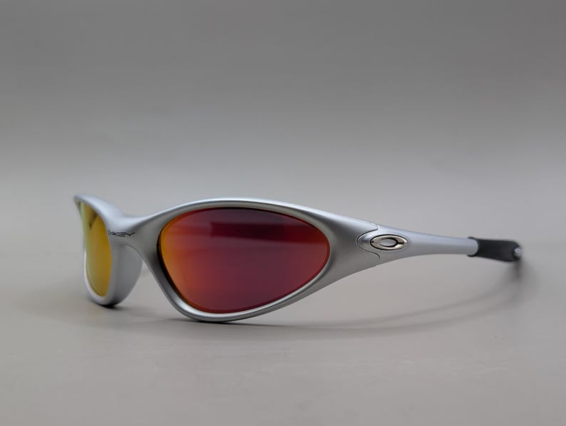 May include: Silver-framed sunglasses with red and yellow lenses. The sporty eyewear features a curved design and black temple tips. The Oakley logo is visible on the frame.