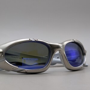 May include: Silver-framed sunglasses with blue lenses. The sunglasses have a futuristic design with a metallic finish. The text "MADE IN U.S.A." is visible on the lens. The arms are white.