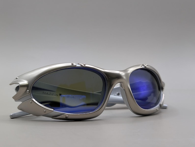 May include: Silver-framed sunglasses with blue lenses. The sunglasses have a futuristic design with a metallic finish. The text "MADE IN U.S.A." is visible on the lens. The arms are white.