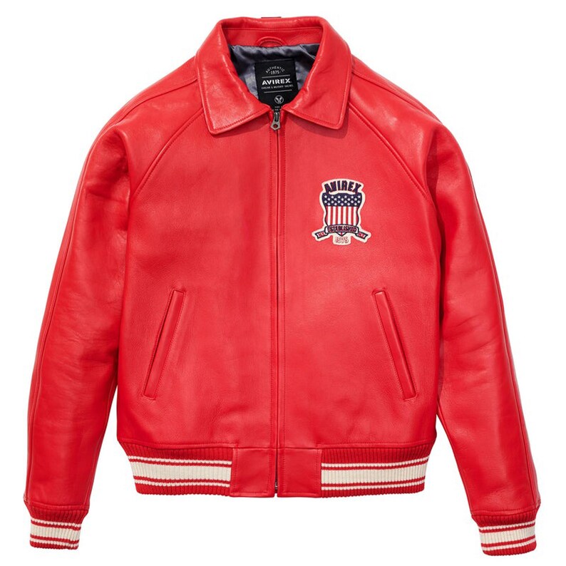 May include: A bright red leather jacket with a zip-up front, collar, and ribbed cuffs and hem. The jacket features a patch with the text "AVIREX" and an American flag design on the chest. The cuffs and hem have white stripes.