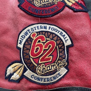 Peut inclure: Gros plan sur une veste en cuir rouge avec des &eacute;cussons brod&eacute;s. Les &eacute;cussons comportent le texte "Midwestern Football 62 Champs Conference" dans un design circulaire avec des accents dor&eacute;s et bleu marine. La veste a une bordure noire.