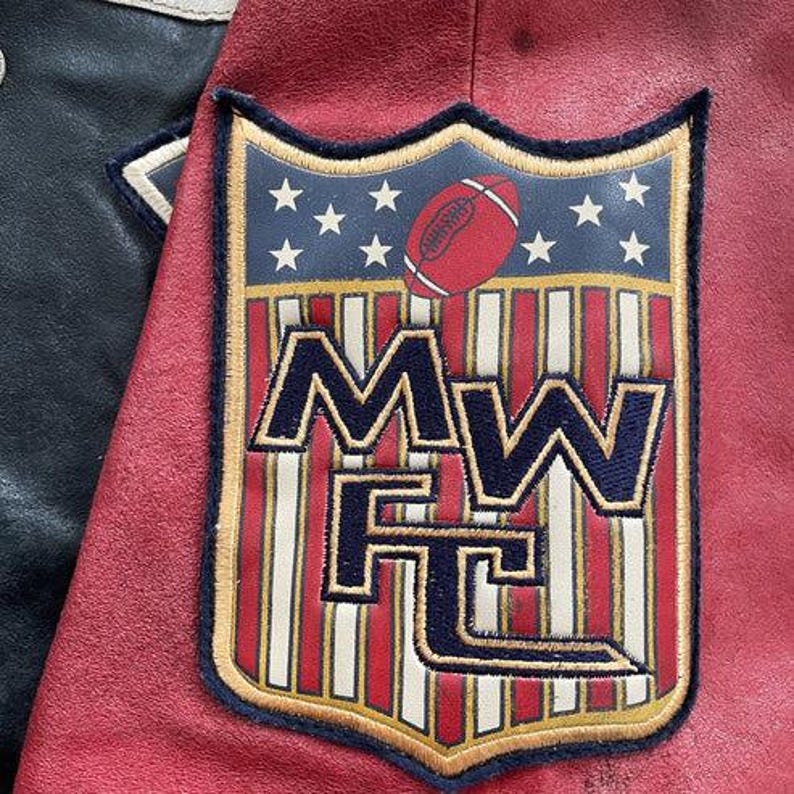 Peut inclure: Gros plan sur une veste universitaire rouge et noire avec un &eacute;cusson sur le th&egrave;me du football. L'&eacute;cusson repr&eacute;sente un ballon de football, des &eacute;toiles et les lettres "MW FC" dans un motif de bouclier. La veste a une texture semblable &agrave; du cuir.