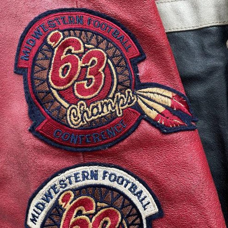 Peut inclure: Gros plan sur une veste en cuir rouge avec des &eacute;cussons brod&eacute;s. Les &eacute;cussons comportent les mots "Midwestern Football" et "63 Champs Conference" dans un motif circulaire. Un motif de plume est &eacute;galement visible.