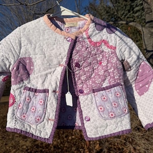 May include: A quilted jacket in white, lavender, and purple, with floral and geometric patterns. The jacket has a round collar, patch pockets, and button closures. The jacket is hanging on a white hanger.