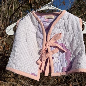 Pode incluir: Um colete acolchoado em tons de branco, lavanda e rosa, pendurado em um cabide branco. O colete apresenta um design de patchwork com fecho de laço e acabamento rosa. O fundo é um ambiente natural ao ar livre com galhos e luz solar.