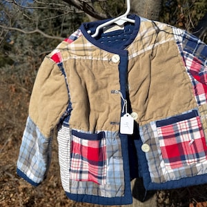 May include: A patchwork jacket with a mix of plaid, corduroy, and solid fabrics in shades of blue, red, tan, and white. The jacket has a round collar, button closures, and two front pockets. It is hanging on a white hanger.