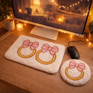 May include: A cream-colored desk mat and coaster set with embroidered pink bows and gold rings. The set includes a rectangular mat and a round coaster, both with a fluffy texture. A black computer mouse is next to the coaster.
