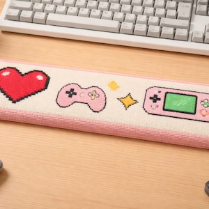 May include: A rectangular desk mat with pixel art designs. It features a red heart, a pink game controller, a yellow star, and a pink handheld console. The mat has a pink border and is on a light wood surface.
