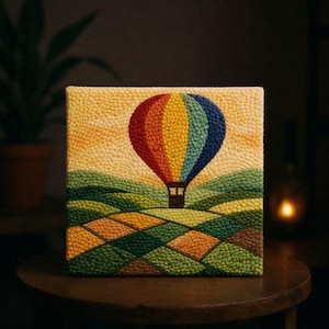 May include: A square textile artwork shows a hot air balloon in flight over a patchwork landscape. The balloon is multi-coloured, with red, orange, yellow, and blue panels. The fields below are green and brown. The background is a pale yellow.