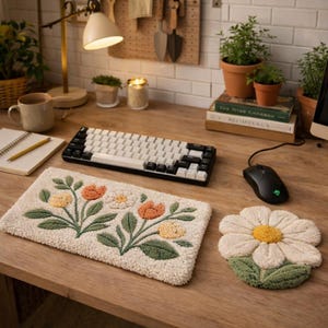 May include: A beige floral-patterned desk mat and a daisy-shaped coaster sit on a wooden desk, alongside a black and white keyboard and a black computer mouse. The desk is lit by a desk lamp and has a mug, notebook, and potted plants.