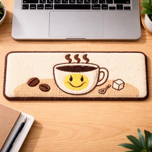 Op de afbeelding: Een rechthoekige beige muismat met een bruine rand toont een lachende koffiekop, koffiebonen, een suikerklontje en een lepel. De koffiekop heeft een gele smiley en stoom die opstijgt. De mat ligt op een houten bureau.