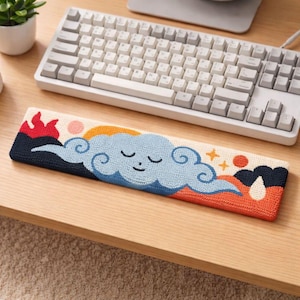 Puede incluir: Apoyamuñecas bordado rectangular con un diseño caprichoso de nube. El apoyamuñecas tiene una nube azul con los ojos cerrados, un sol naranja y llamas rojas. Se coloca delante de un teclado blanco sobre un escritorio de madera. Mide unos 30 cm.