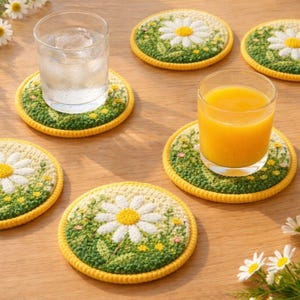 Puede incluir: Un conjunto de posavasos redondos hechos a mano con un diseño de flor de margarita. Cada posavasos presenta una margarita blanca con un centro amarillo, rodeada de detalles bordados en verde y amarillo. Vasos de agua y zumo.