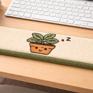 May include: A rectangular beige mouse mat with a green border. The centre features a cartoon image of a potted plant with green leaves and a smiling face. The letter "z" is next to the plant.