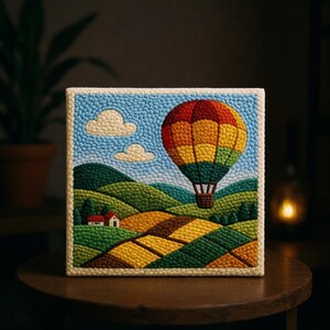 May include: A square textile artwork showing a hot air balloon in flight above a patchwork landscape. The balloon is a vibrant mix of red, orange, yellow, and green. The scene includes hills, fields, clouds, and a small house, all set against a blue sky, with a cream border.