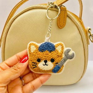 以下が含まれることがあります： 青い帽子と灰色のレンチを持った、手作りのオレンジ色の猫のキーホルダー。 猫は黒い目と白い口を持っています。 キーホルダーは、シルバーチェーンと丸いキーリングに付いています。