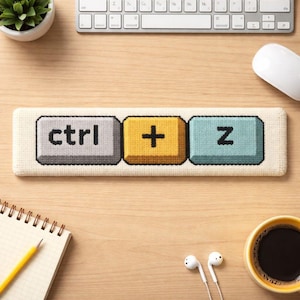May include: A beige, rectangular keyboard wrist rest featuring embroidered keyboard keys: "ctrl", "+", and "z". The wrist rest is on a wooden desk, with a keyboard, mouse, notepad, and coffee cup in the background.