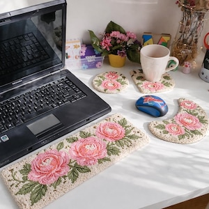 May include: A collection of handmade floral-themed desk accessories. Includes a rectangular mousepad, coasters, and a crescent-shaped mat, all featuring embroidered pink roses and green leaves on a beige background. A laptop, mug, and flowers are also present.