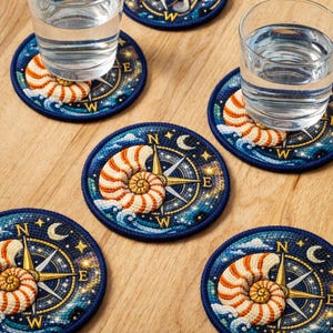 Op de afbeelding: Ronde onderzetters met een nautisch thema. Elke onderzetter heeft een kompasontwerp met een zeeschelp en een sterrenhemel in blauw, goud en wit. De onderzetters zijn gemaakt van een textuurrijk materiaal en beschermen oppervlakken tegen waterkringen.