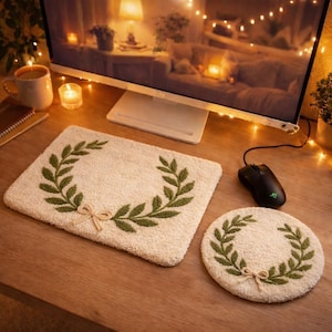 Op de afbeelding: Een crèmekleurige rechthoekige muismat en een ronde onderzetter, beide met een groen geborduurd laurierkransontwerp en een kleine strik. De items liggen op een houten bureau, naast een zwarte computermuis.