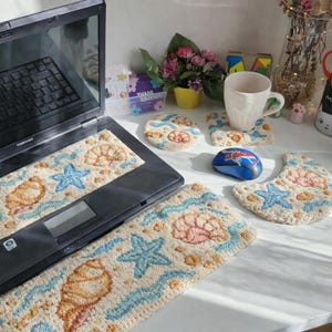 May include: A laptop with a seashell-themed rug and matching coasters. The rug features a beige background with blue waves, starfish, and seashell designs. A blue mouse and a white mug are also on the desk. The overall theme is coastal and beach-inspired.