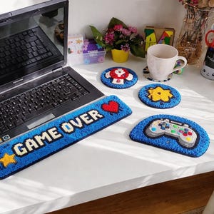 Puede incluir: Colección de artículos hechos a mano con temática de videojuegos en azul y blanco. Incluye un reposamuñecas para teclado "GAME OVER", tres posavasos redondos con diseños de videojuegos y un posavasos con forma de mando.