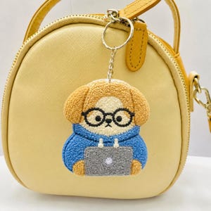 May include: A dog-shaped keychain with a blue hoodie, glasses, and a laptop. The keychain is attached to a yellow purse with a gold zipper and strap. The dog has brown fur and is wearing black glasses.