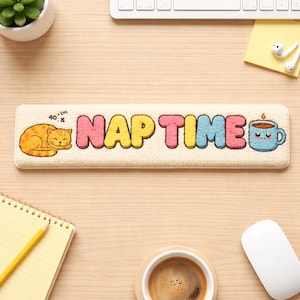 Könnte beinhalten: Eine rechteckige, beige Schreibtischunterlage mit den Worten "NAP TIME" in bunten Buchstaben. Links ist eine orange Katze eingerollt, rechts eine blaue Kaffeetasse. Die Matte misst 40 cm x 8 cm.