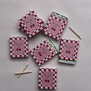 May include: A collection of decorative matchboxes with a cherry design and the text "Mon chéri". The matchboxes are red with a white heart pattern border. Several matchsticks are visible.