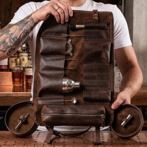May include: A brown leather bartender tool roll with multiple pockets and compartments, holding various bar tools. The roll is open, displaying a shaker, strainer, and other stainless steel tools. The roll is held up by a person.