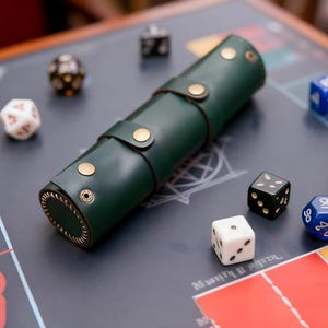 May include: A dark green leather dice roll with brass-colored snaps and a stitched edge, surrounded by various dice on a game board. Dice include white, black, red, and blue, with different numbers of sides.