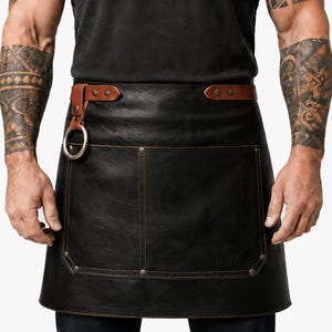 May include: A black waist apron featuring a smooth texture, a brown leather waistband with metallic rivets, and a large front pocket. A silver-toned metal ring is attached to the left side of the waistband with a brown leather strap.
