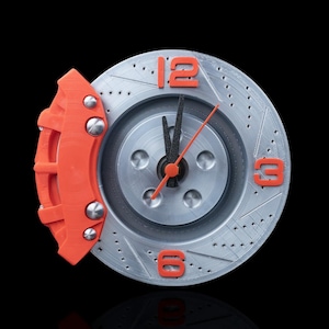 Peut inclure: Une horloge argentée avec un design de rotor de frein de voiture. L'horloge comporte des chiffres orange à 12, 3 et 6 heures, et un étrier de frein orange. Les aiguilles sont noires et orange. L'horloge est sur fond noir.