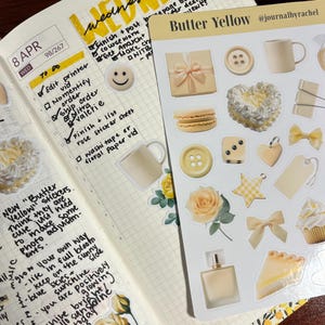 May include: A planner spread with handwritten notes and a sticker sheet labeled "Butter Yellow." The stickers include images of a gift box, buttons, a mug, a heart, a bow, a star, a perfume bottle, a macaron, a rose, and a slice of pie.