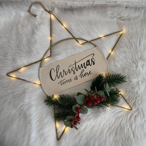 Może przedstawiać: Złota metalowa dekoracja świąteczna w kształcie gwiazdy z ciepłymi białymi diodami LED. Baner z napisem "Christmas time is here" jest wyśrodkowany, z sztuczną zielenią i czerwonymi jagodami na dole. Do zawieszenia przymocowany jest sznurek jutowy.
