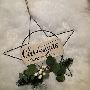 Może przedstawiać: Czarna metalowa gwiazda świąteczna z drewnianym znakiem z napisem "Christmas time is here". Ozdobiona zielonymi liśćmi, białymi jagodami i sznurkowym wieszakiem.