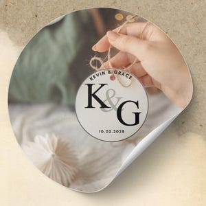 Könnte beinhalten: Ein runder Aufkleber mit den Namen "KEVIN & GRACE" und den Initialen "K & G", sowie dem Datum "10.02.2028". Der Aufkleber wird an einer Schnur gehalten. Der Hintergrund ist unscharf.