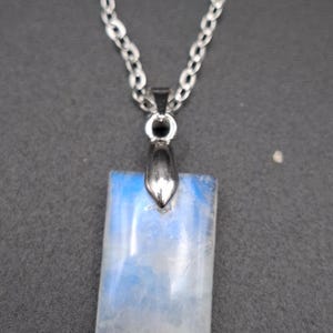 Peut inclure: Un collier argenté avec un pendentif rectangulaire en pierre irisée bleue et blanche. Le pendentif est suspendu à une chaîne délicate. Le bijou est présenté sur un fond gris foncé, mettant en valeur ses couleurs.