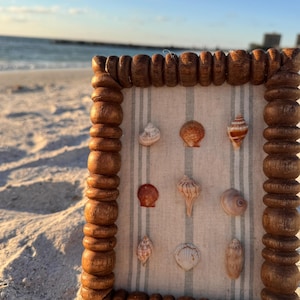 May include: A wooden framed display of seashells on a sandy beach. The frame has a decorative, turned-wood design. The seashells are various shapes and colours, arranged on a striped fabric background.