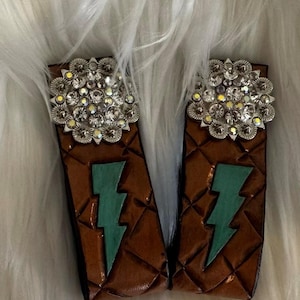 May include: Pair of brown leather cuffs with a quilted pattern and a turquoise lightning bolt design. Each cuff features a silver-toned, flower-shaped embellishment with clear rhinestones. The cuffs are displayed on a white, fluffy surface.