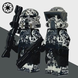 May include: Two minifigure toys of soldiers in camouflage armor, holding black rifles. The helmets and armor feature a gray, white, and black camouflage pattern. The Imperial symbol is in the upper left corner.