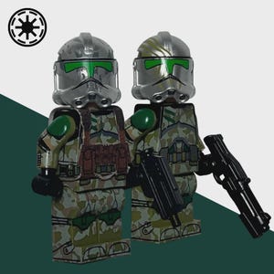 May include: Two toy soldiers in camouflage armor with silver helmets and green visors. They are holding black weapons. The soldiers are standing in front of a white and green background. A black and white symbol is in the upper left corner.