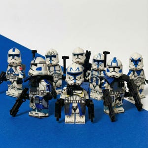 May include: A group of Lego Star Wars Clone Troopers, with white and blue armour, helmets, and blasters. The figures are displayed against a blue and white backdrop, highlighting the detailed design.
