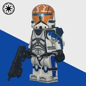 Op de afbeelding: Een Lego-minifiguur van een Star Wars-cloon-trooper. De figuur is voornamelijk wit met oranje en blauwe accenten, en een zwarte blaster. De helm heeft oranje markeringen. De achtergrond is wit en blauw met een zwart-wit symbool.