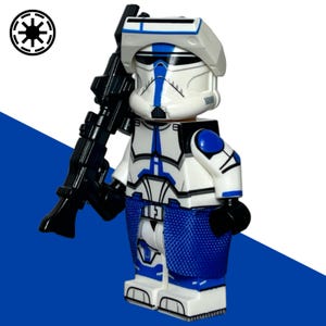 May include: A white and blue plastic toy soldier minifigure wearing a helmet with a visor, holding a black blaster rifle. The figure features detailed armor markings in blue and black, with a blue fabric kama skirt around the waist and a black Galactic Republic symbol in the corner.