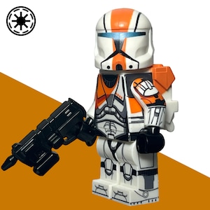 Könnte beinhalten: Eine weiße und orangefarbene Spielzeugsoldaten-Minifigur mit einem Helm mit blauem Visier, schwarzen Details und einem passenden orangefarbenen Rucksack. Die Figur hält eine schwarze Blaster-Waffe aus Kunststoff und zeigt ein kreisförmiges schwarz-weißes Emblem.